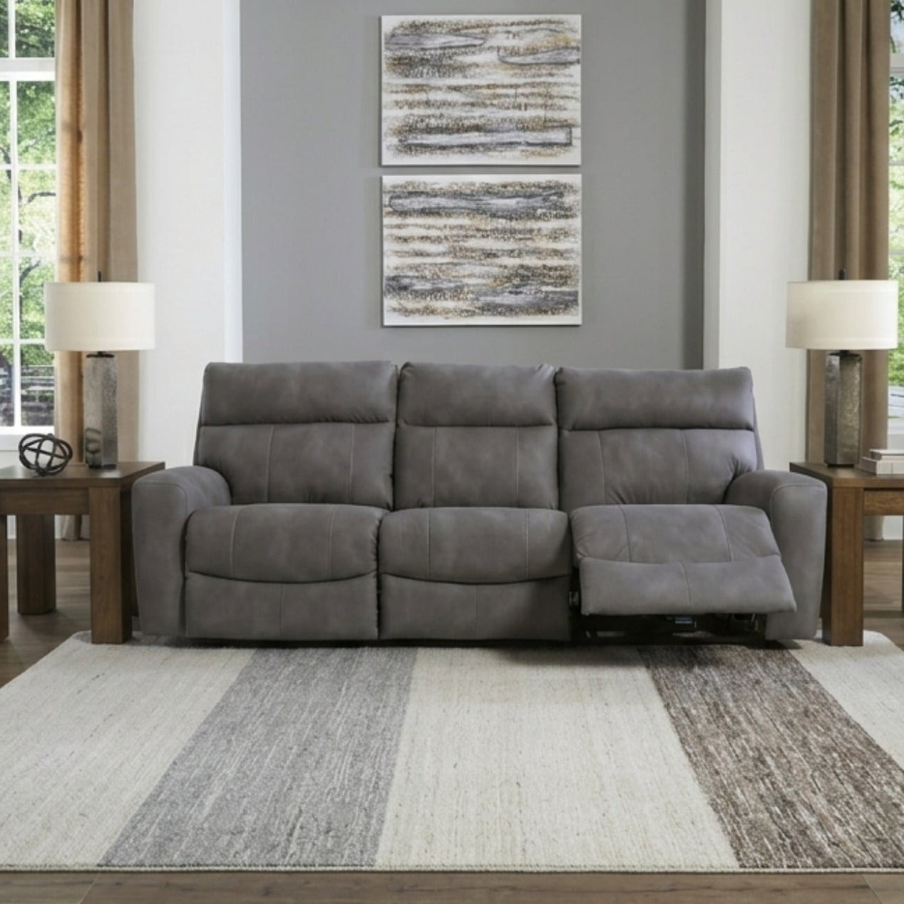 A gray three-seat recliner sofa in a modern living room setting with wood floors, beige curtains, and a striped area rug.
