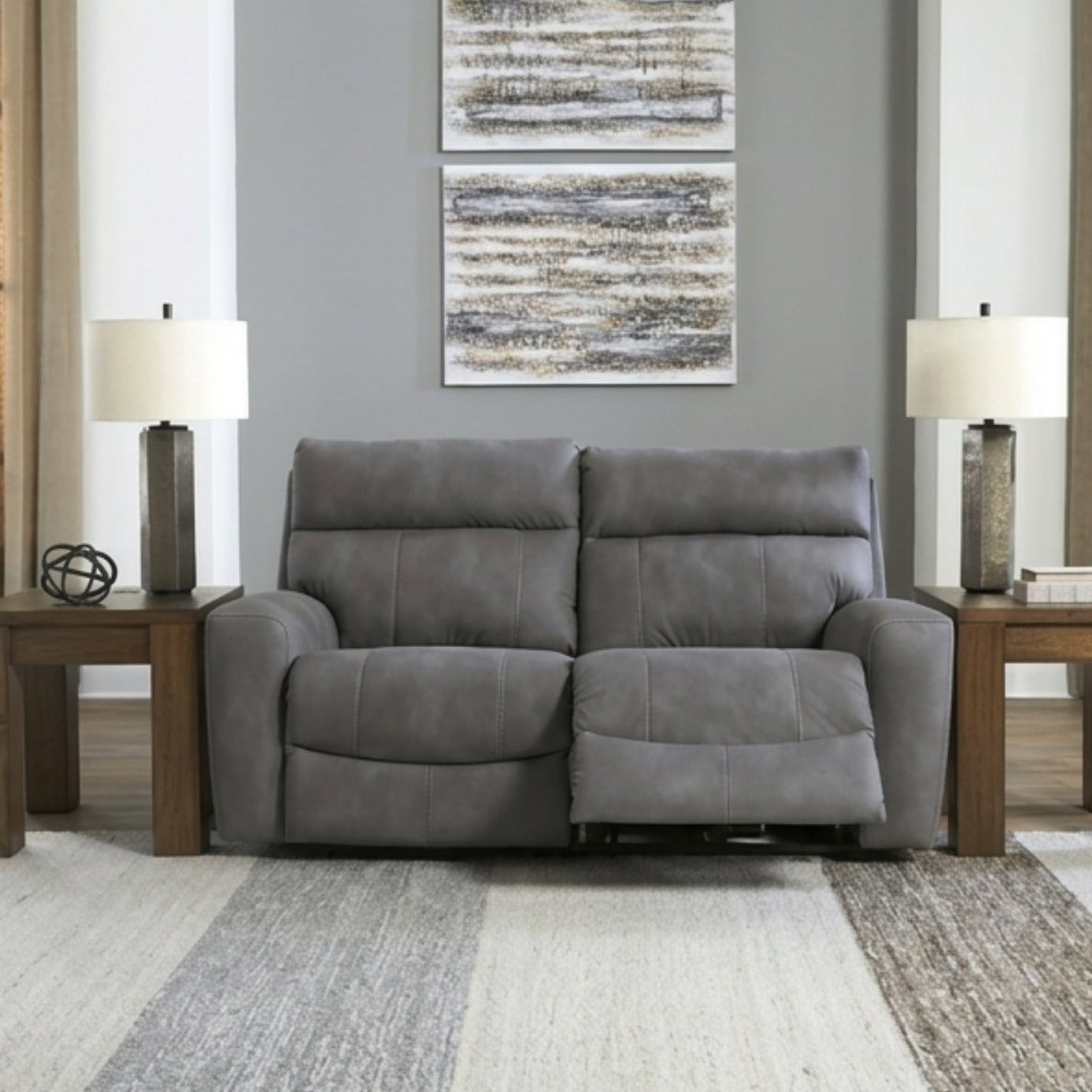 A gray upholstered reclining loveseat centered in a staged living room with two wooden side tables, matching table lamps, a striped area rug, and wall art above the sofa.