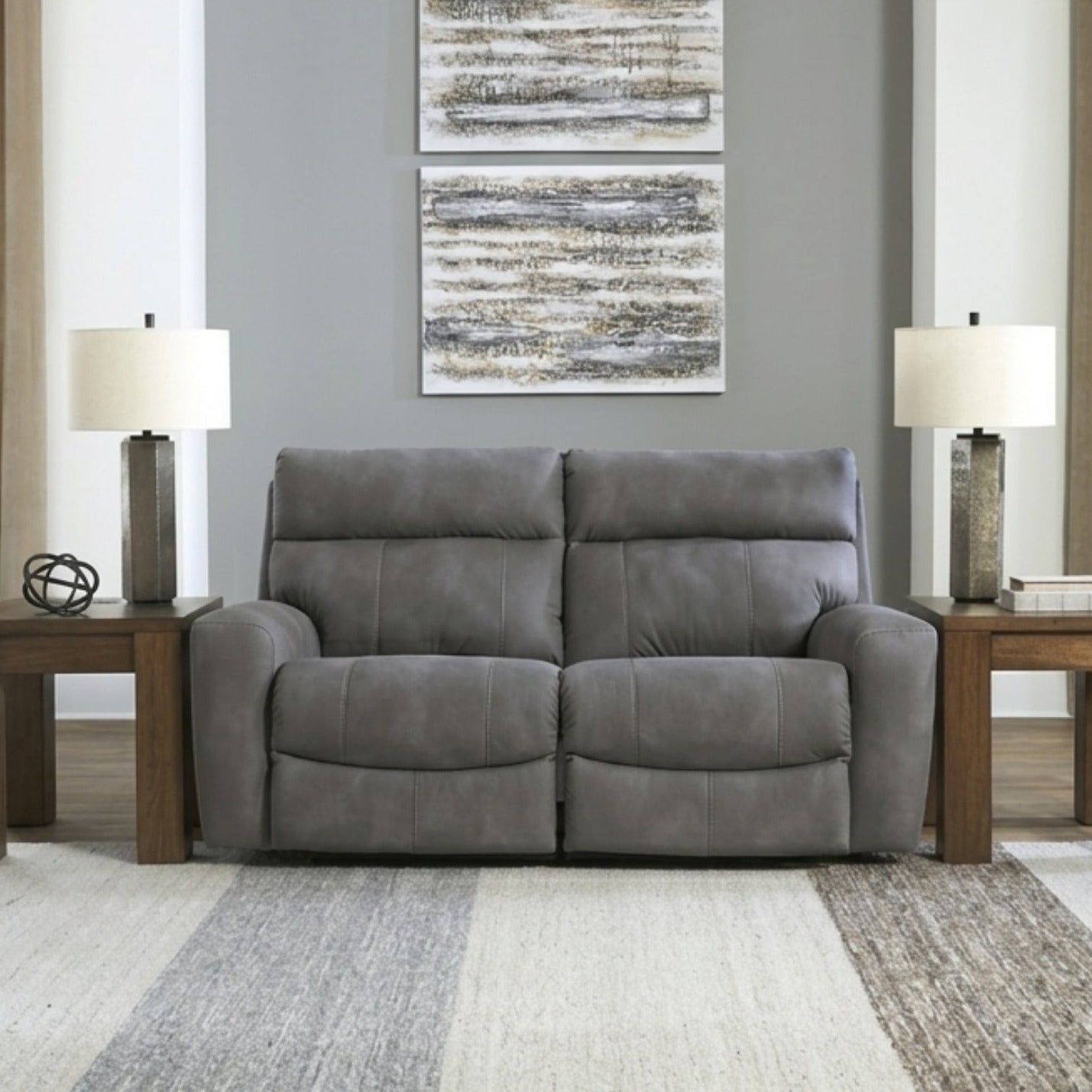 A gray two-seater reclining loveseat sofa centered in a bright living room, flanked by matching wooden side tables with table lamps, a patterned area rug, and two windows with beige curtains.