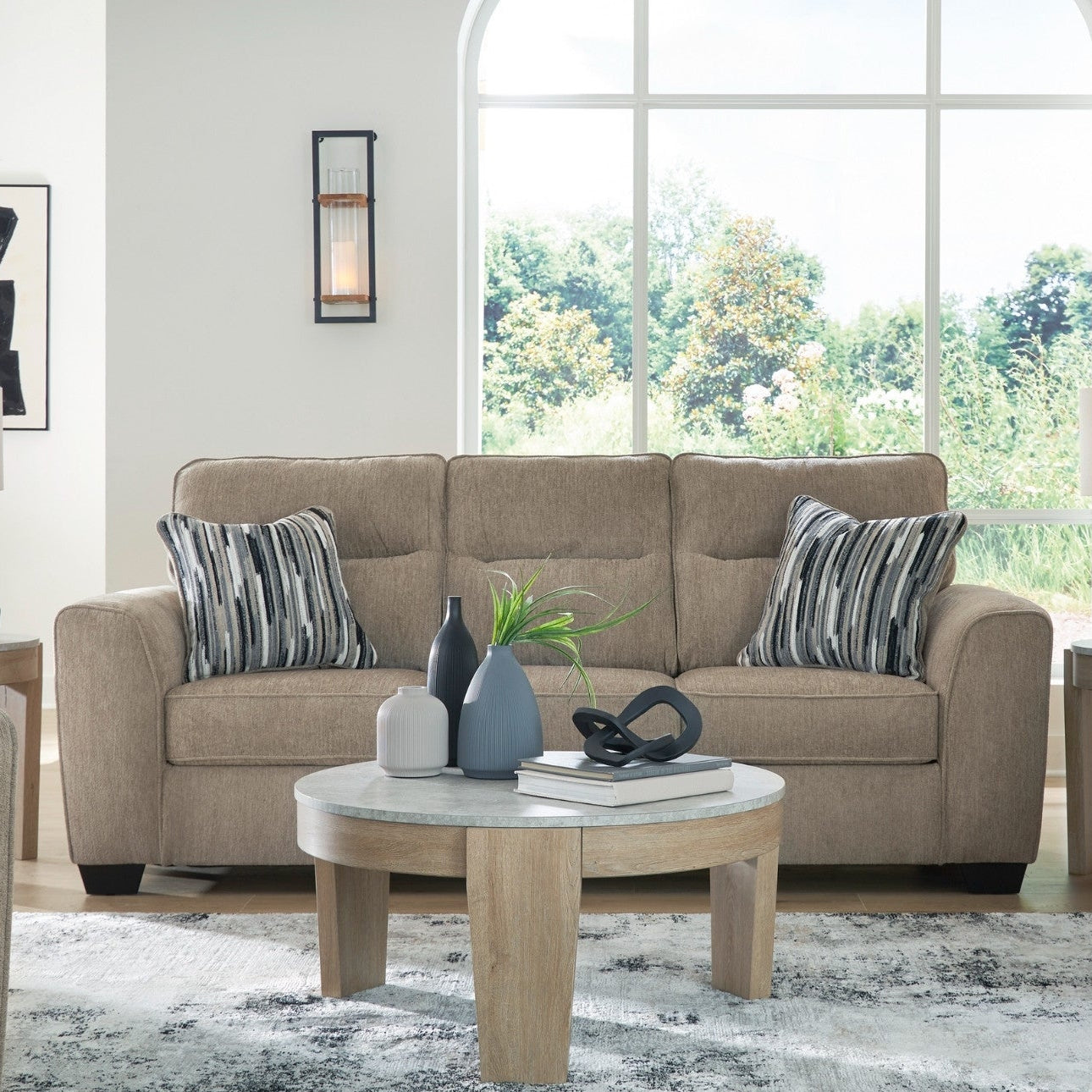 A staged living room showing a beige upholstered three-seat sofa, matching loveseat, striped throw pillows, a round light wood coffee table, side tables, lamps, wall sconces, and a patterned rug in front of a large arched window.